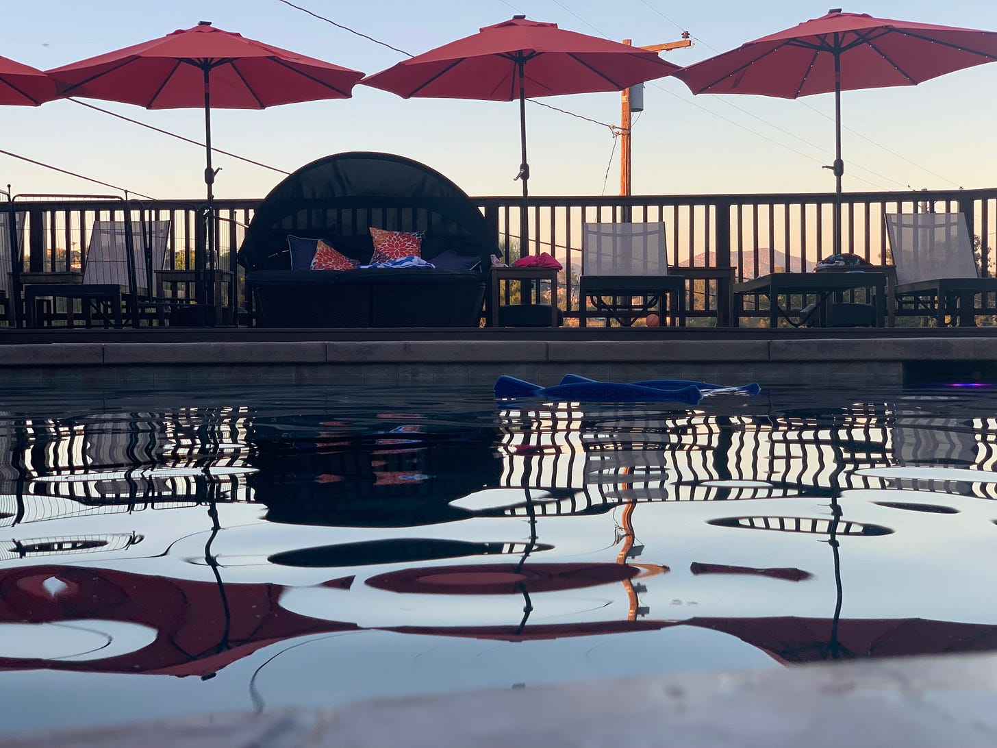 A pool at dusk, surrounded by beach chairs and orange umbrellas, reflected in the water.