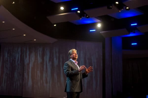 A man in a suit with no tie speaks on a stage with his hands raised.