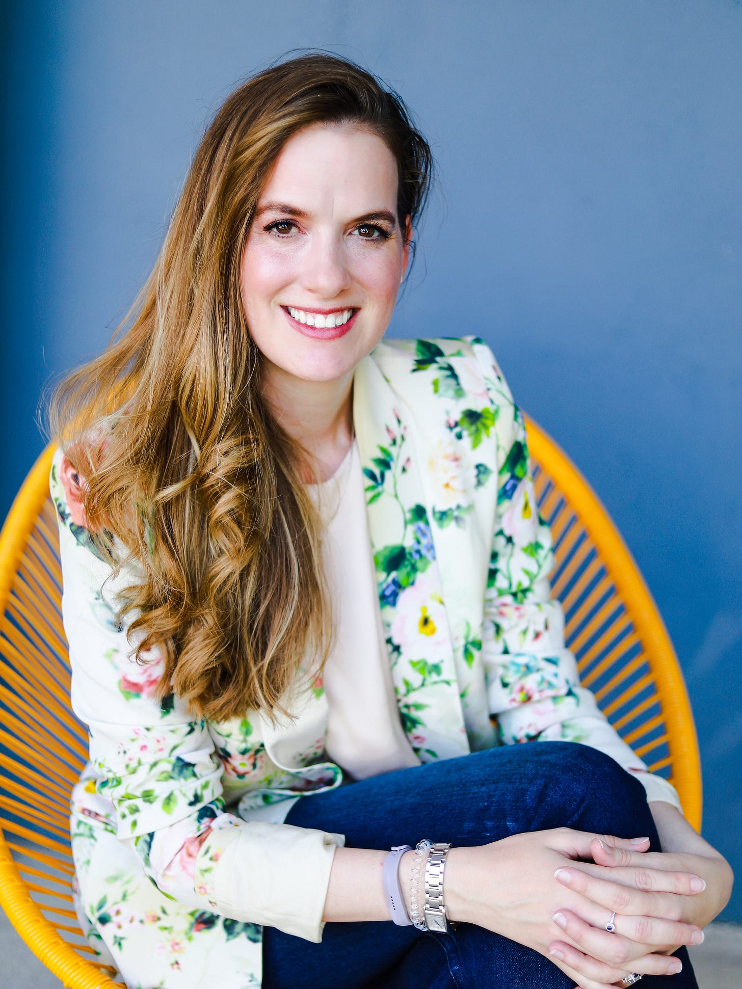 A profile photo of Jennifer Millspaugh. She is sitting in a yellow chair against a vibrant blue wall. She is wearing a cream blazer with flowers and blue jeans.