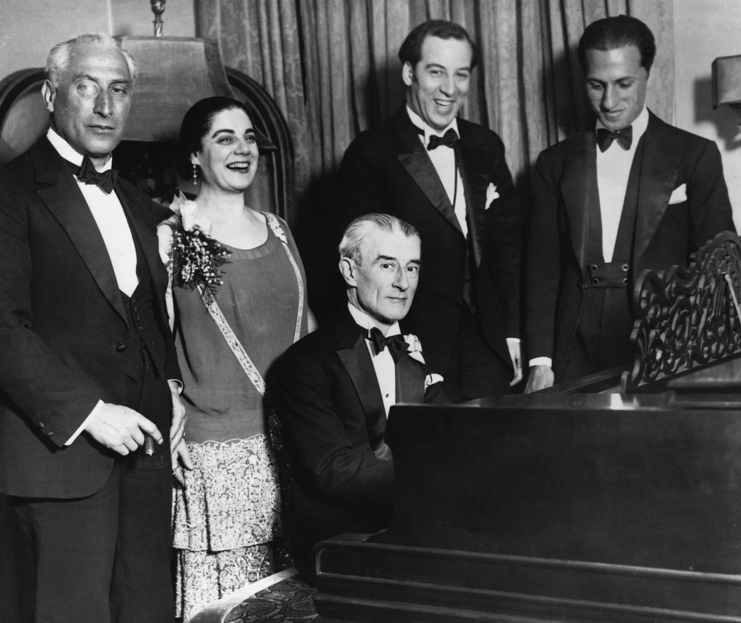 Black and white photograph of four men in tuxedos and one woman in an elegant party dress standing around a grand piano Black and white photograph of four men in tuxedos and one woman in an elegant party dress standing around a grand piano