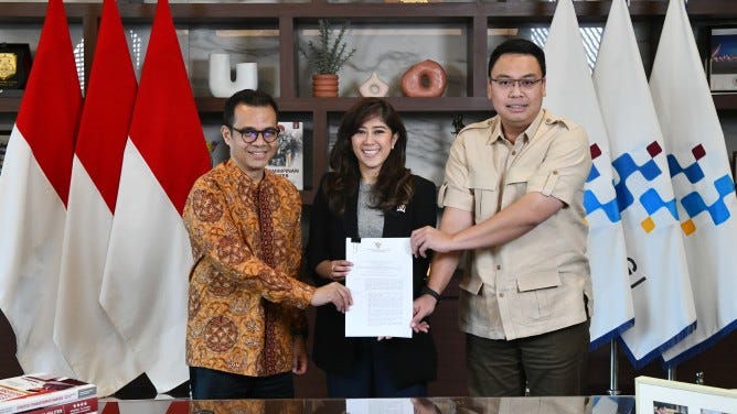 A medium wide full shot of three people standing behind a desk in an office. On the left, a man wearing glasses and an orange and brown batik shirt holds a document with both hands. In the centre, a woman with long brown hair wearing a black blazer and grey shirt stands behind the desk, smiling. On the right, a taller man wearing glasses and a tan short-sleeved button-down shirt holds the same document with both hands. Behind them on the left are three Indonesian flags. Behind them on the right are three white flags with a blue, yellow, and red pixelated logo. In the background is a wooden shelf with several decorative items, including a white vase and two orange ceramic rings.