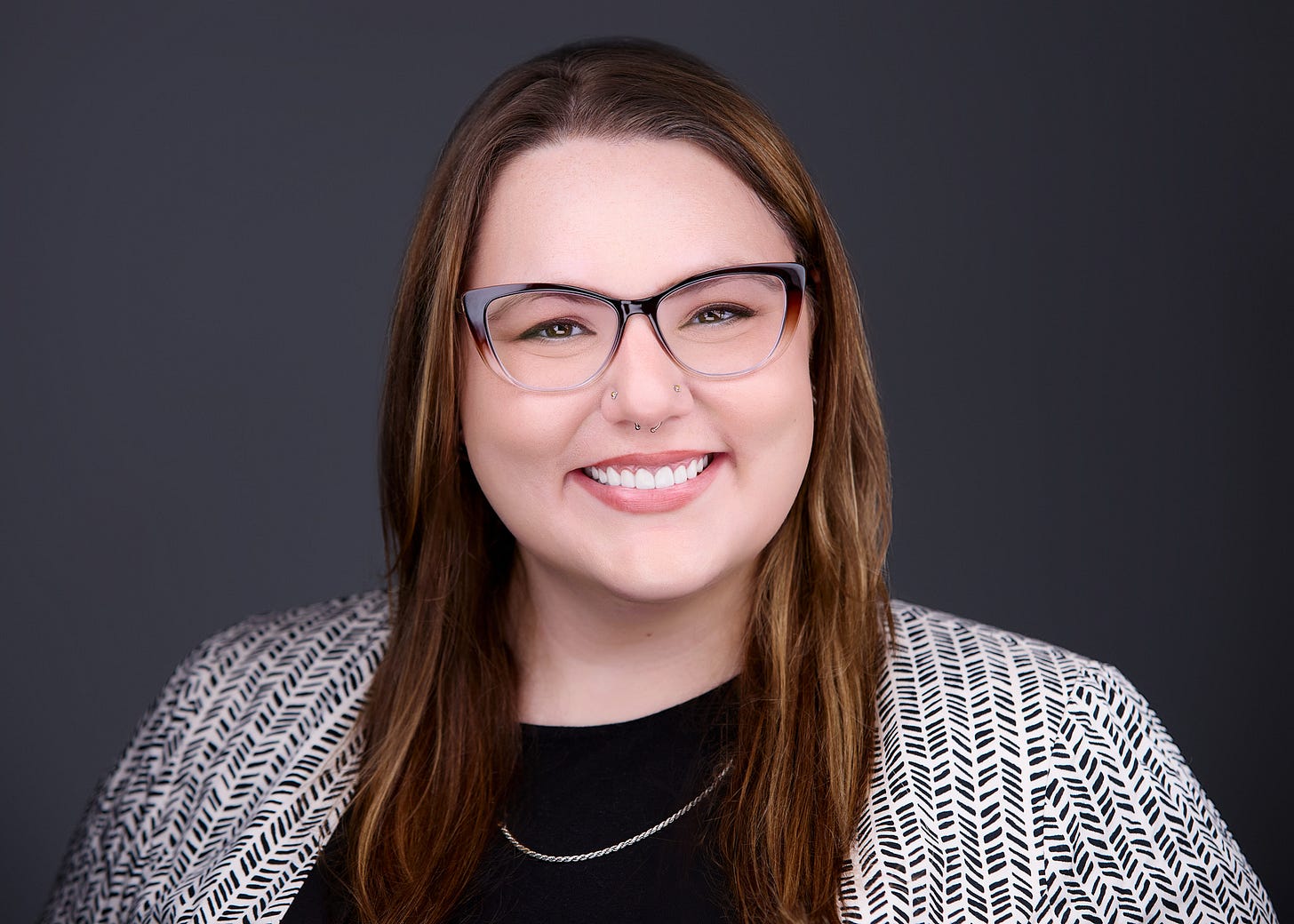 A studio portrait shows a person with long brown hair, glasses, and facial piercings, smiling directly at the camera. They wear a black top and a patterned blazer, with soft lighting that creates gentle contrast against a dark, gradient background. The overall composition feels contemporary and professional, conveying warmth and confidence.