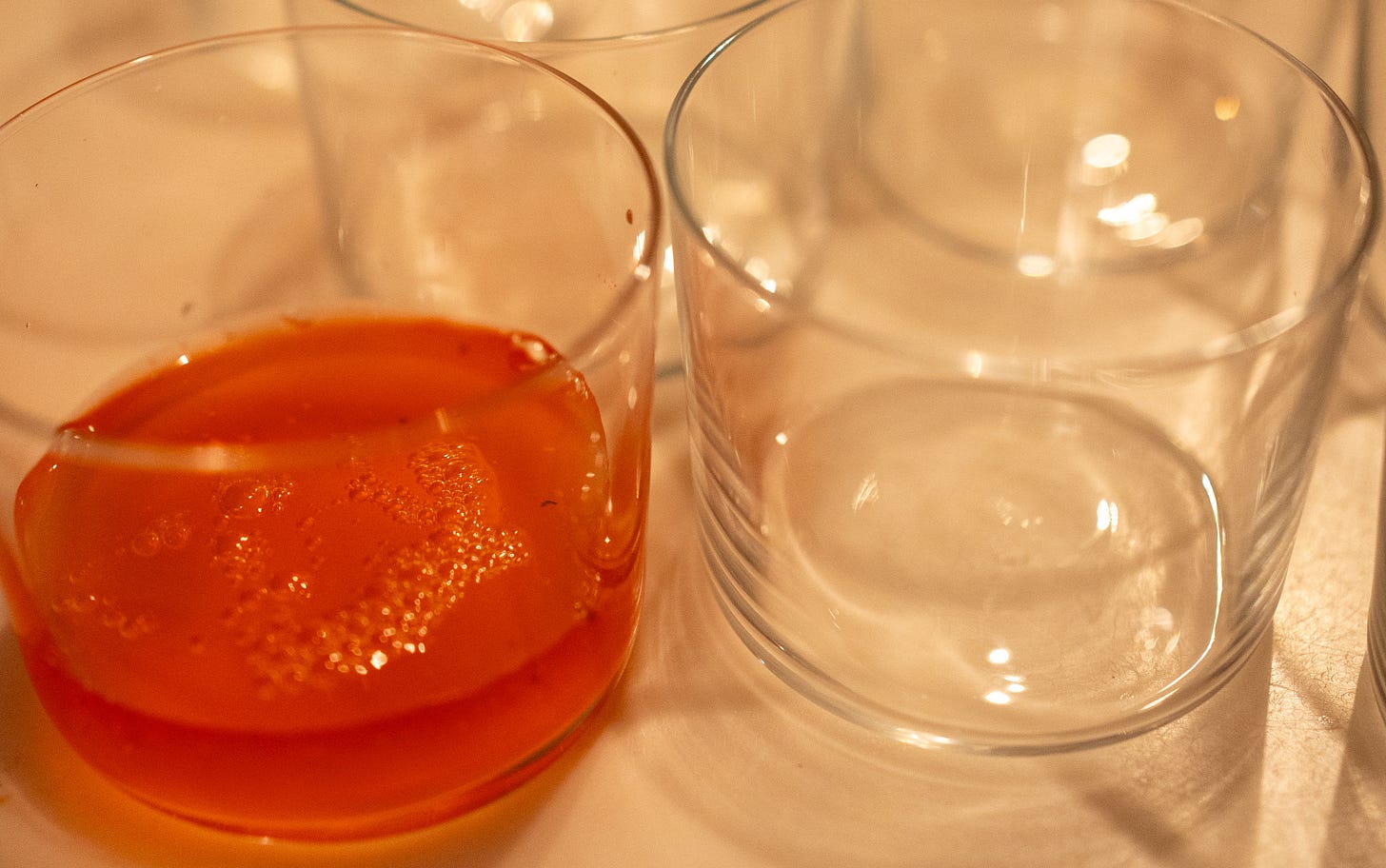 Close up of clear drinking glasses on a white surface, with one in the foreground filled with a bright reddish-orange carbonated liquid.