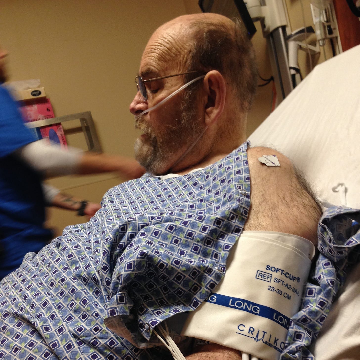 An elderly bearded man with glasses in a blue hospital gown and a blood pressure cuff, his eyes are closed.