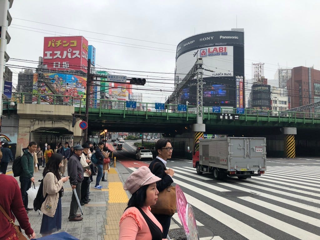 Many people wait to cross a wide pedestrian crossing while a light truck passes by. A rail bridge is in the background and tall buildings with advertising on them are behind that. Many people wait to cross a wide pedestrian crossing while a light truck passes by. A rail bridge is in the background and tall buildings with advertising on them are behind that.
