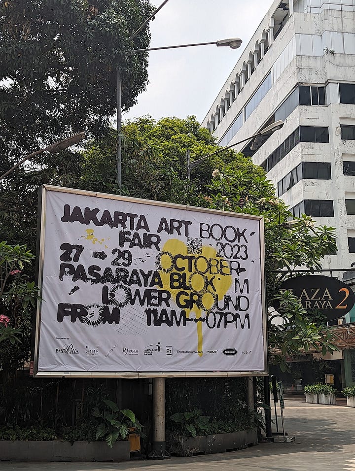The first image is alarge outdoor banner advertising the Jakarta Art Book Fair 2023. The banner displays details such as the dates (27-29 October 2023), location (Pasaraya Blok M, Lower Ground), and hours (11 AM - 7 PM). The banner is surrounded by greenery with a tall white building visible in the background. The second image A busy scene at a booth during the Jakarta Art Book Fair 2023, with a group of people gathered around a table displaying books and stickers. Several attendees are browsing through the books and taking photos, while the table is covered with colorful books, sticker packs, and promotional materials.
