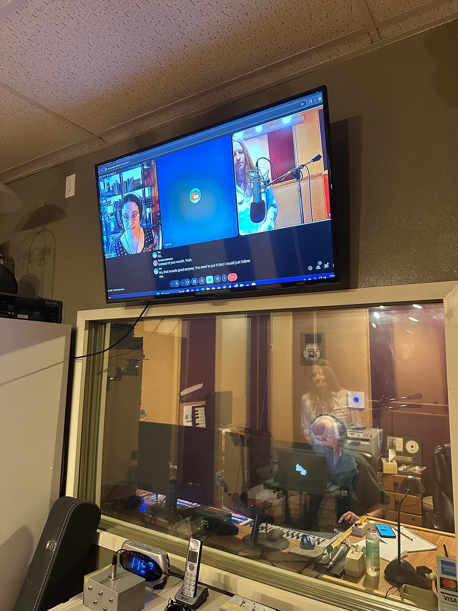 Photo of my view inside the recording studio: gazing at a glass window with a sound engineer sitting beyond, plus a computer screen set up above featuring Google Meet video call and auto-captions.