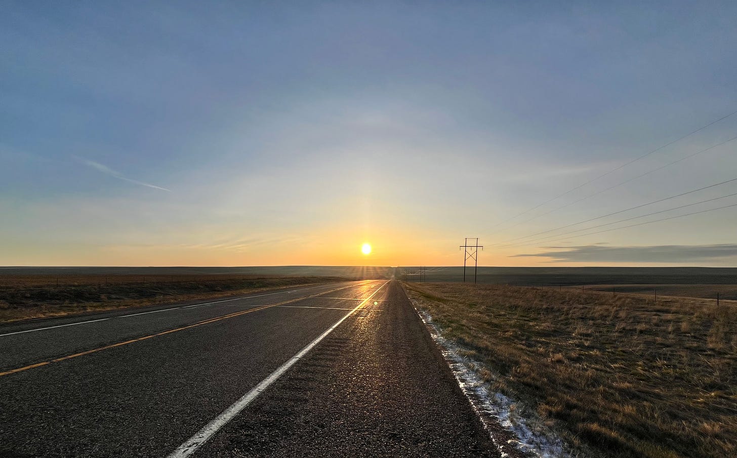 A road in the middle of nowhere with flatlands all around and a sunrise on the horizon