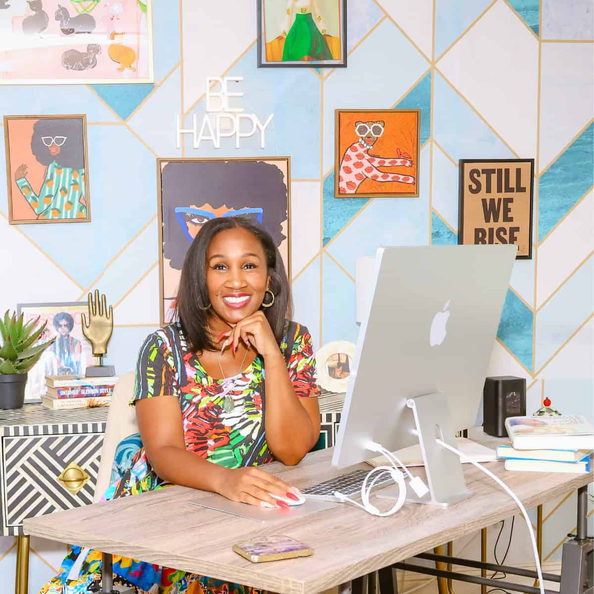 Shaunda Necole smiling at her desk in a colorful home office, working at her iMac computer with bold artwork, motivational quotes, and vibrant decor in the background. A bright, modern workspace that reflects her personality as a soul food expert, Las Vegas insider, author, and digital creator.