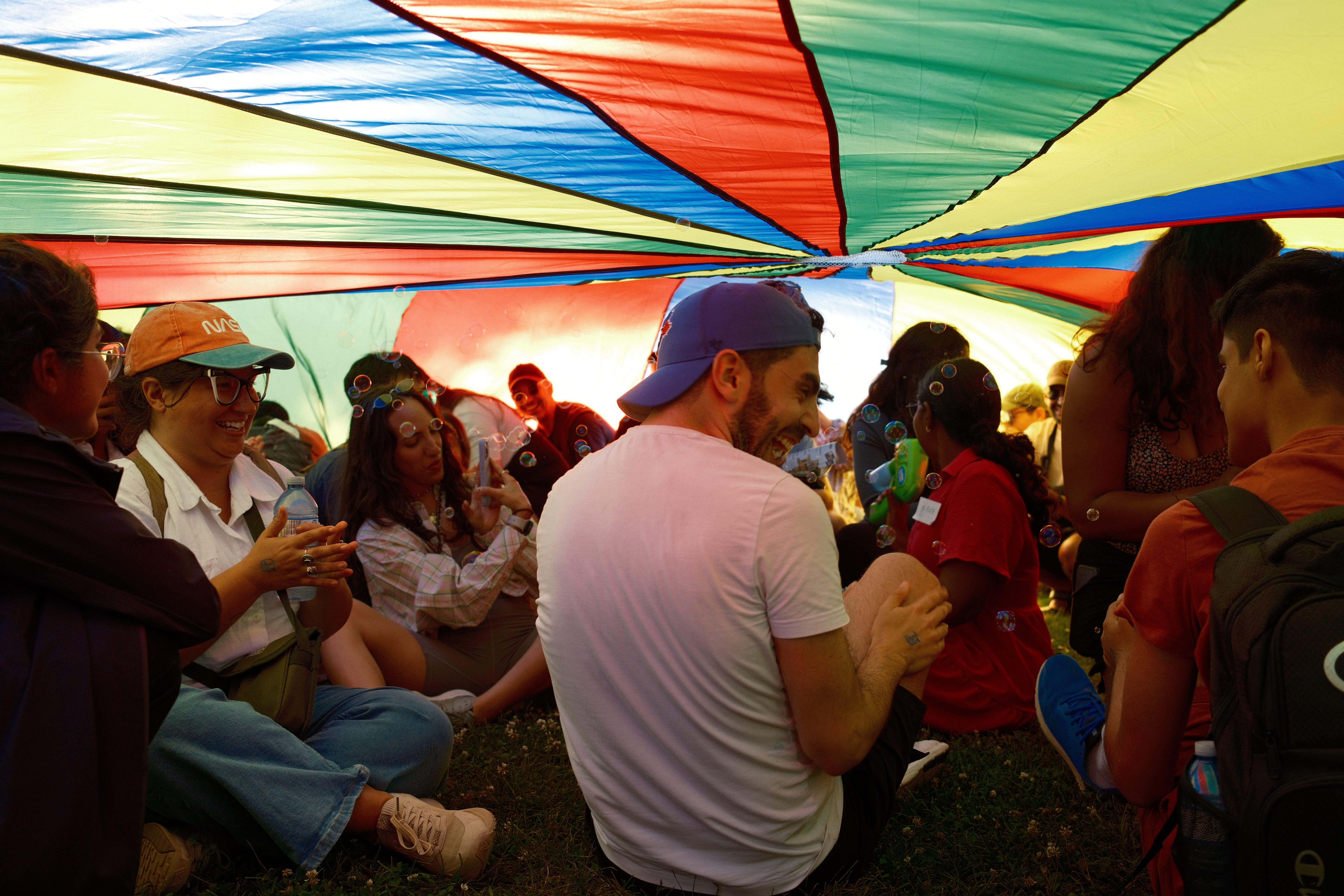 A group of people, including Matthew, smiling and blowing bubbles under a rainbow parachute.