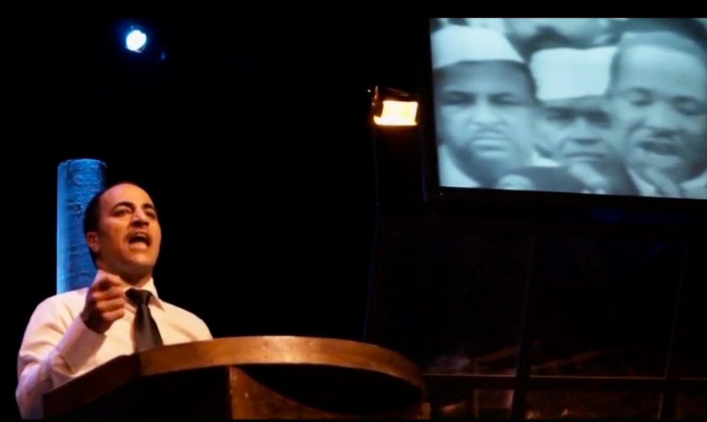  Image from the play "Passages of Martin Luther King." The Palestinian actor who plays King stands at a podium to the left, speaking. Behind him is a large screen with the actual video footage of Dr. King at the podium during the 1963 March on Washington.