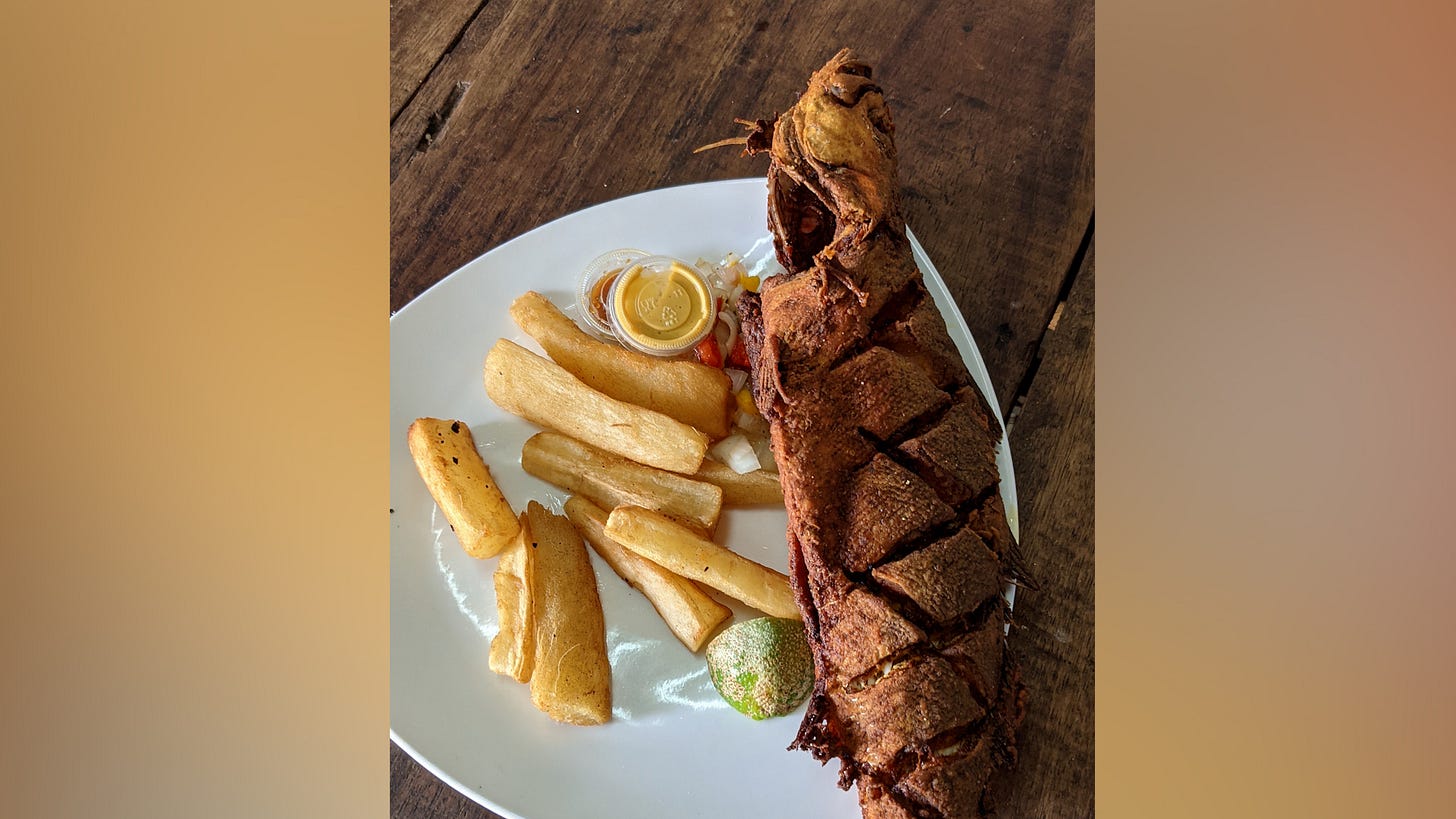 A white triangular plate holds cassava fries and a whole fried fish. A wedge of lime and condiments are also on the plate.