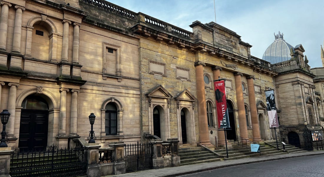 A brown stone building with arched windows and a black fence, with a grey path in front of it A brown stone building with arched windows and a black fence, with a grey path in front of it