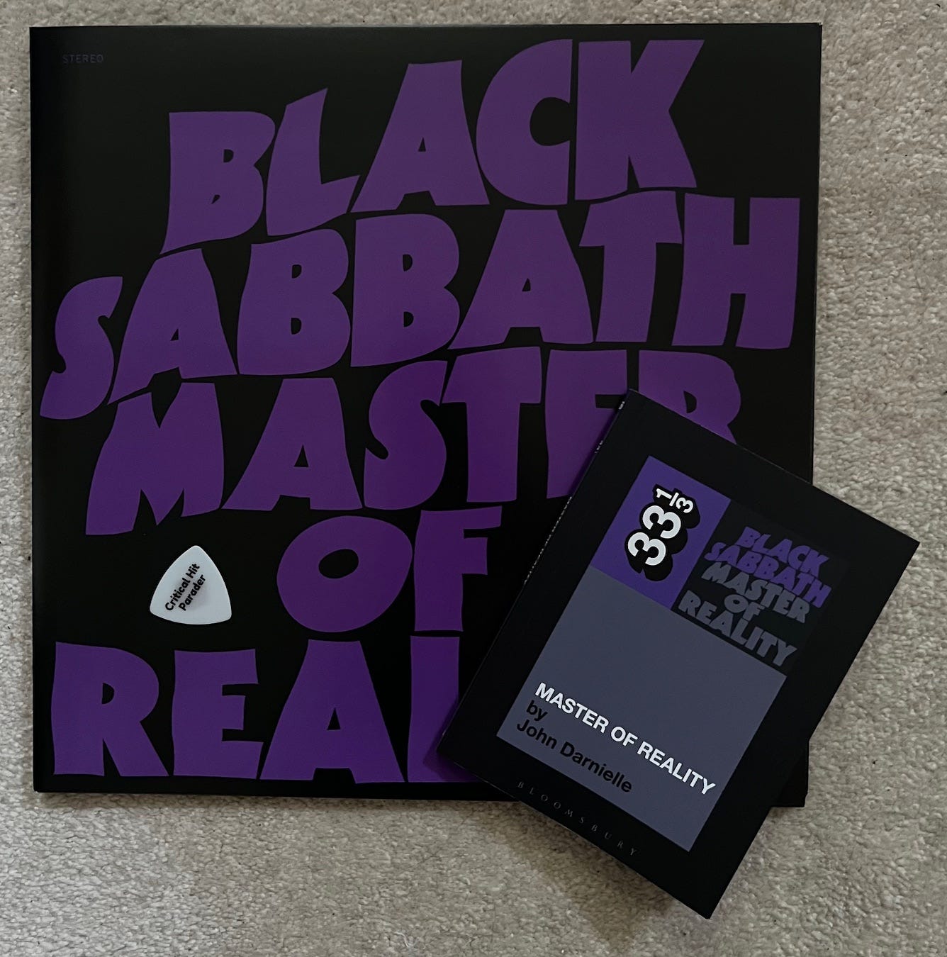 A photo of Black Sabbath's Master of Reality album. It is black with a purple title filling the album cover. A small paperback book of John Darnielle's 33 1/3 book about the album lies on top of the bottom right corner of the record. A white Critical Hit Parader guitar pick sits like an Orchid on top of the album.