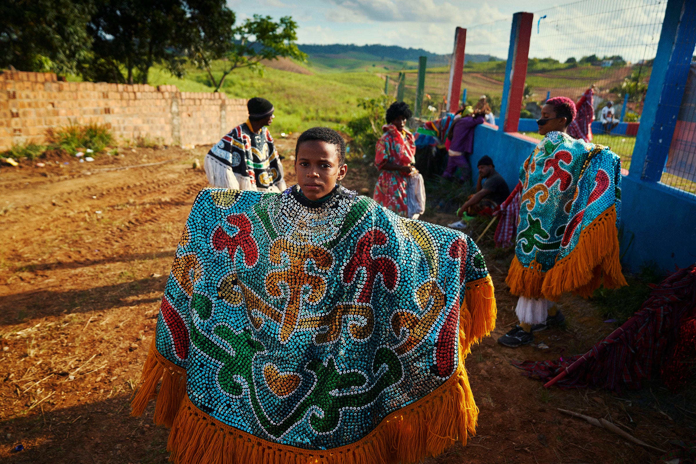 A young person in a brightly colored, beaded costume with fringe stands outdoors while others in similar festive attire gather in the background near a fence and grassy field.
