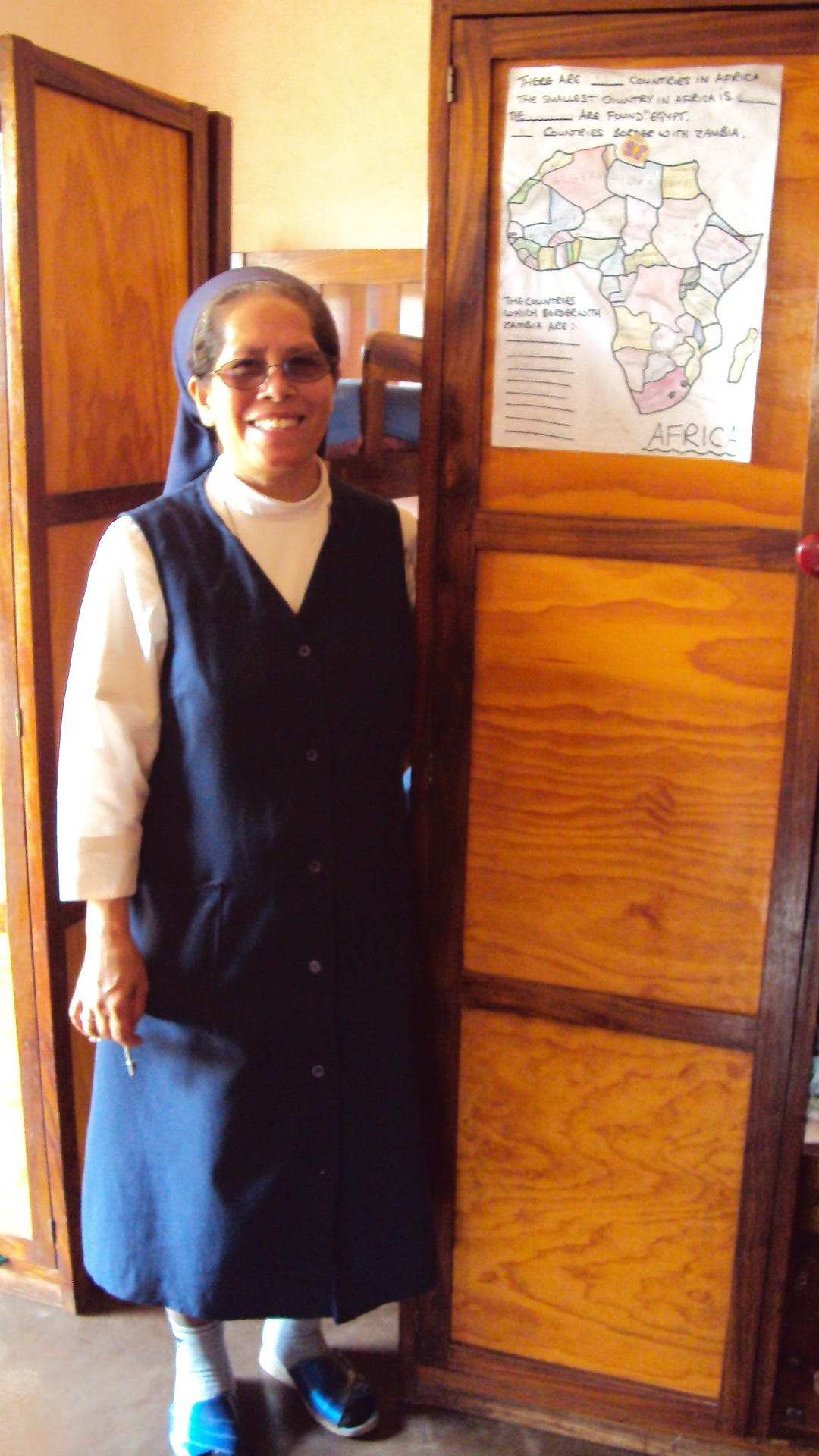 A Black nun wearing a navy-blue habit and sunglasses is smiling. She is standing in front of a cabinet and hand-drawn map of Africa. 