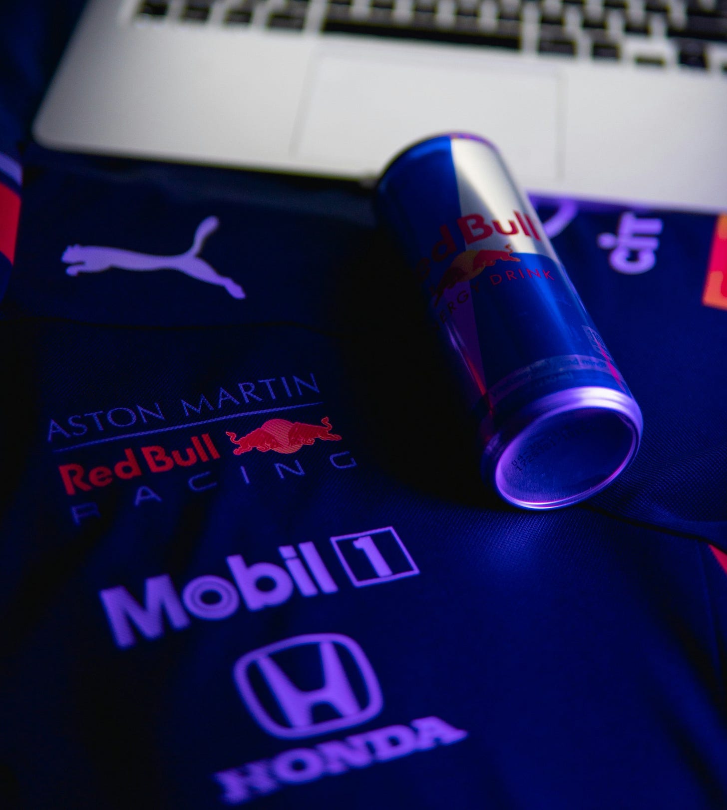 A can of red bull resting on a Red Bull Racing team shirt with an open laptop in the background. The image has blue tinge as if slightly mournful, or haunted, or maybe just modern.