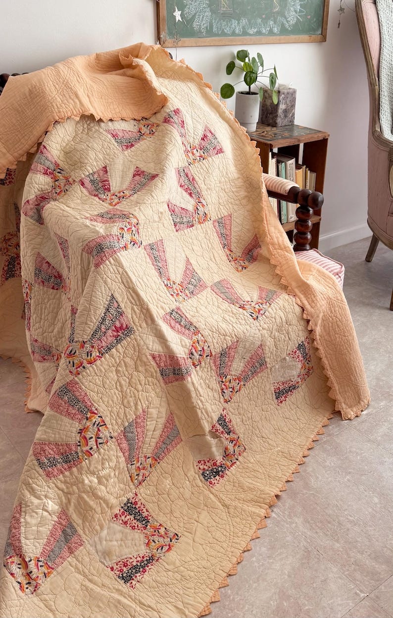 May include: A vintage quilt draped over a chair. The quilt features a cream-colored background with fan-shaped patterns in shades of pink, red, and black. The edges are scalloped and trimmed with a peach-colored fabric. The quilt is a cozy, handmade item. May include: A vintage quilt draped over a chair. The quilt features a cream-colored background with fan-shaped patterns in shades of pink, red, and black. The edges are scalloped and trimmed with a peach-colored fabric. The quilt is a cozy, handmade item.