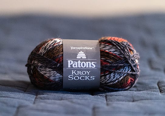 A skein of multicolored yarn sits on a blue blanket. The yarn's label says Patons Kroy Socks