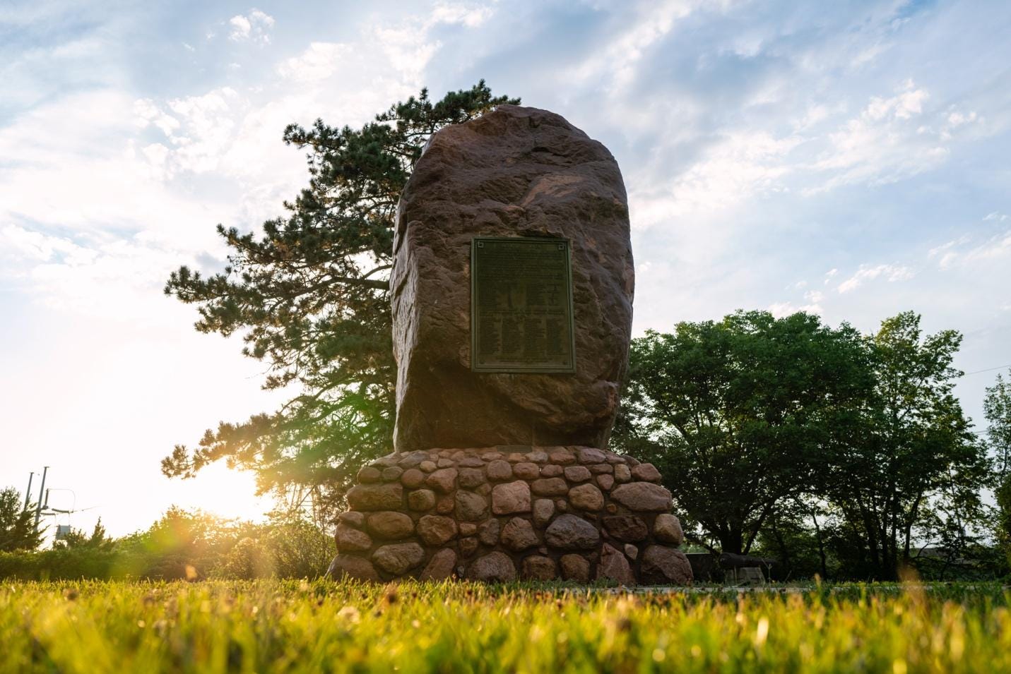 In a clearing at sunrise, a tall red stone boulder sits on a pedestal made of smaller stones. The boulder is almost as tall as the trees around it. 