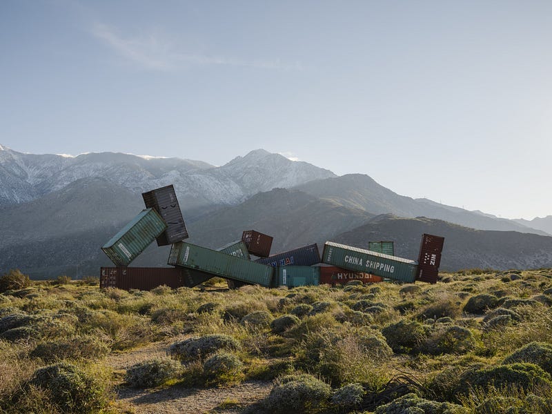 Snow-covered mountains in the distance. A group of massive shipping containers are arranged in the shape of a reclining figure. Its legs are crossed and the head is propped up by one bent arm. Snow-covered mountains in the distance. A group of massive shipping containers are arranged in the shape of a reclining figure. Its legs are crossed and the head is propped up by one bent arm.