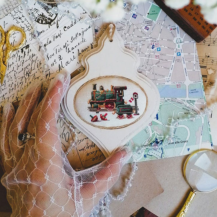 A photo of a gloved hand holding an intricate cross-stitch. It is surrounded by an elaborate, "old timey" scene. A photo of a gloved hand holding an intricate cross-stitch. It is surrounded by an elaborate, "old timey" scene.