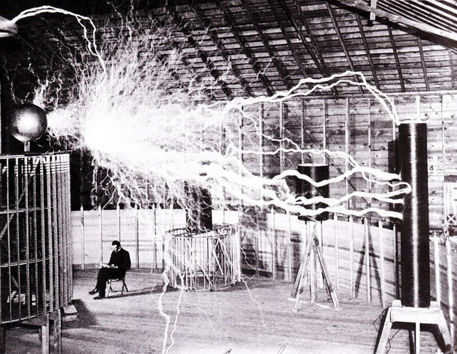 A historical photograph of Nikola Tesla seated calmly in his Colorado Springs laboratory in 1899, surrounded by arcs of white electrical discharge from a massive coil — representing the visible geometry of vibration and resonance at the heart of his experiments.