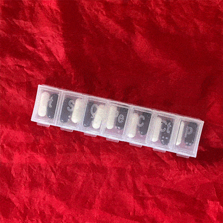 Rainbow Squared Year 5, Piece Twenty-Eight: 7. Red Black White by Rachel Cassandra. A stop-motion photo animation loop of a red background with a clear plastic pill organizer on top of it with seven slots for the days of the week filled with black pills and white pills, where the lids dance open and shut and the pills dance out of the containers and then back into the containers.