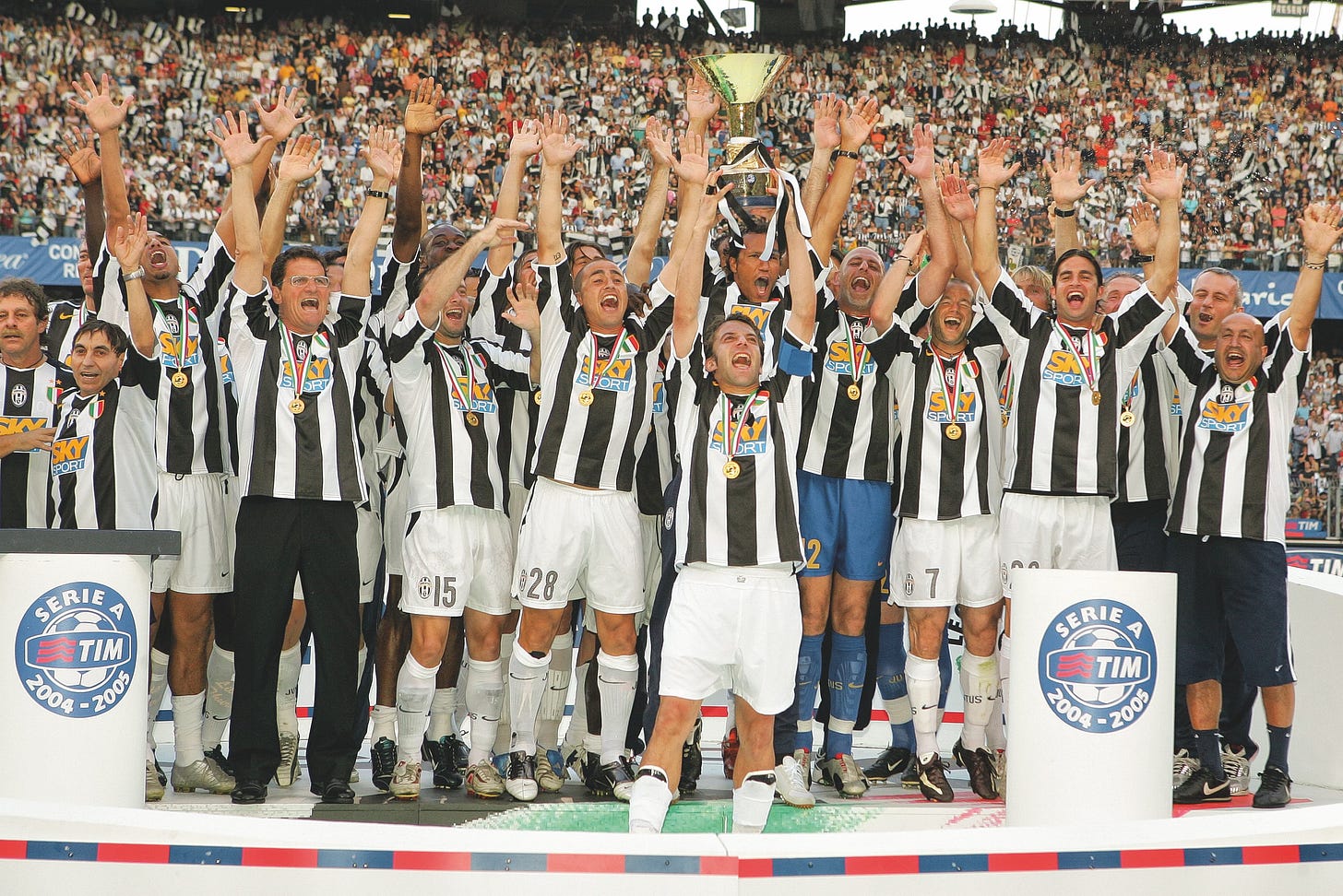 Juventus players and staff in black-and-white kits celebrate with raised arms and the Serie A trophy after winning the 2004–05 title, wearing gold medals on the podium.