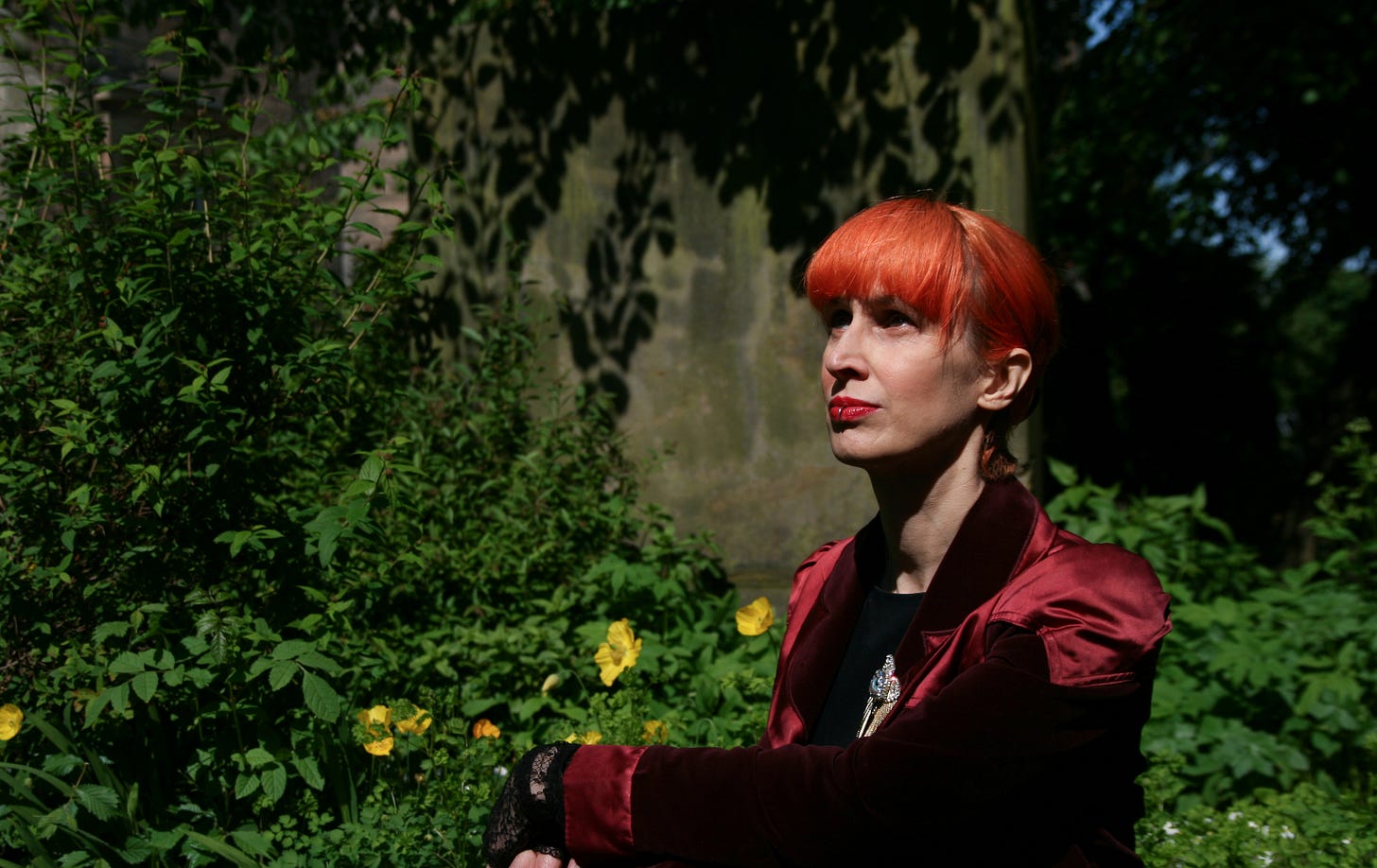 * A thin white woman is basking in the sun in front of greenery and a smattering of yellow flowers. She’s looking off into the distance, towards the left, with a contemplative air. She’s wearing a deep wine velvet coat with silk panels at the shoulders and cuffs, and a gold brooch is just visible. She has bright orange hair pinned up and a long straight fringe, red lipstick, and a silver lip ring. 