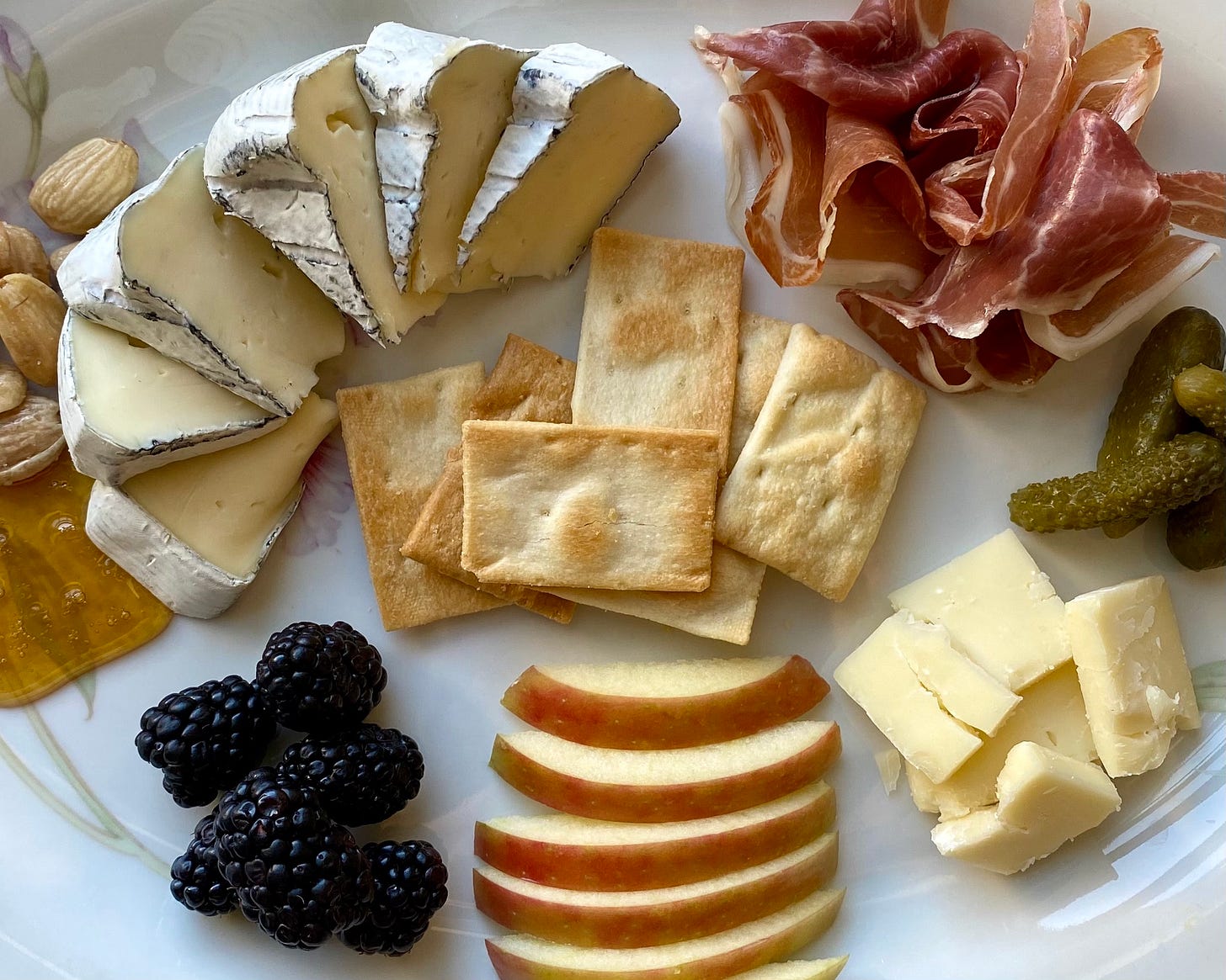 cheese plate with (clockwise from top left) marcona almonds, sliced brie-style cheese, prosciutto, cornichons, prairie breeze cheddar pieces, sliced apple, blackberries, honey, surrounding olive oil crackers in the center cheese plate with (clockwise from top left) marcona almonds, sliced brie-style cheese, prosciutto, cornichons, prairie breeze cheddar pieces, sliced apple, blackberries, honey, surrounding olive oil crackers in the center