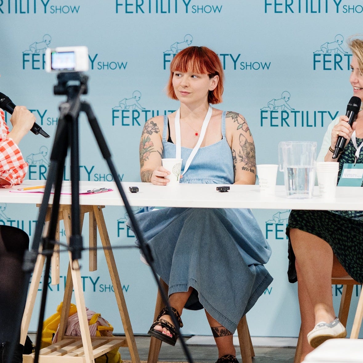 Photograph of @thequeermama at The Ferility Show, sat at a table holding a drink with people holding mircrophones close by