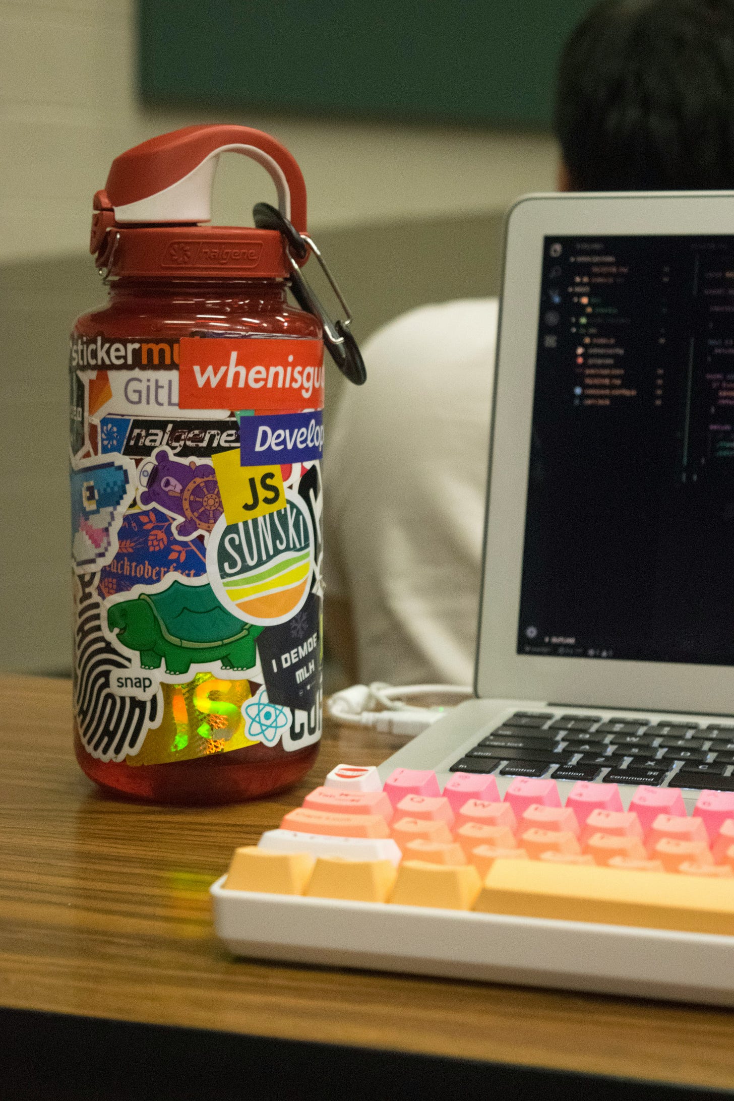 Metal water bottle covered in stickers sitting on a desk to a compter with a multicolored keyboard