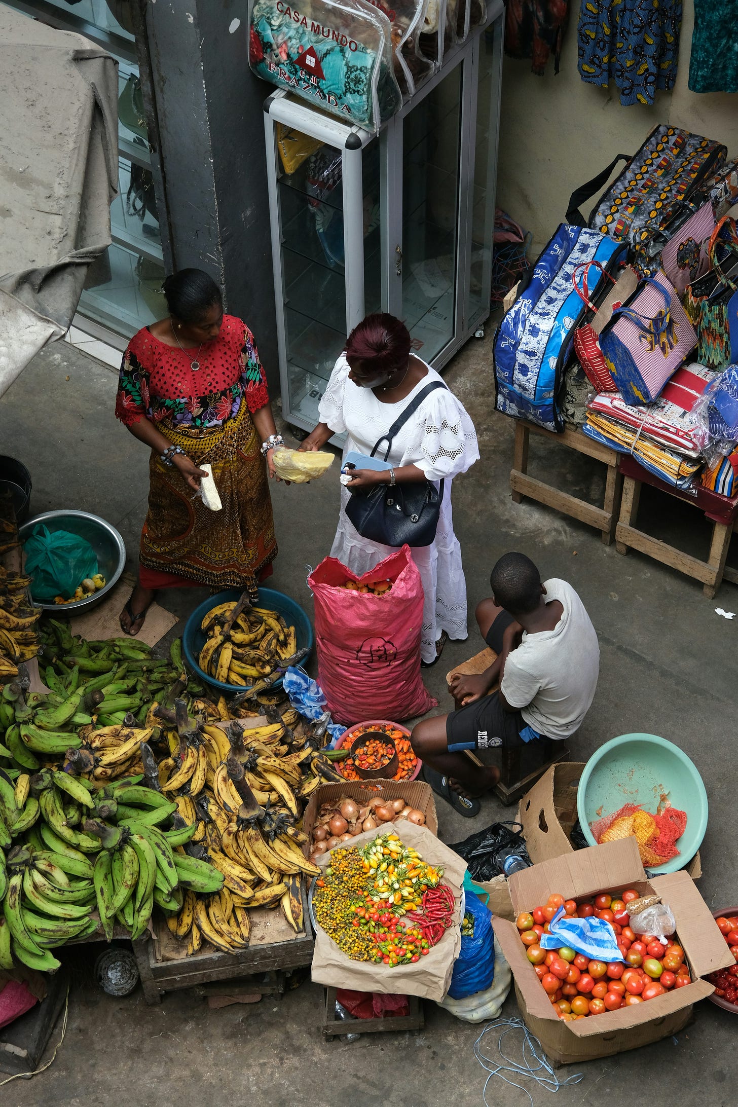 Birds-eye image of two women performing a transaction for plantain at a market. A younger man sits on a stool in front of them, and bowls overflow with yellow and green plantain.