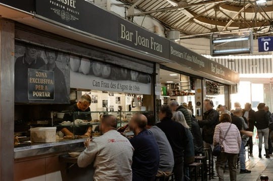 “Interior of Bar Joan Frau in Santa Catalina Market, Palma de Mallorca, with people seated at the counter, bar staff serving food, and a bustling atmosphere.”