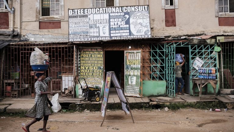 A photo of a woman carrying goods on her head, walking passed a row of shops.