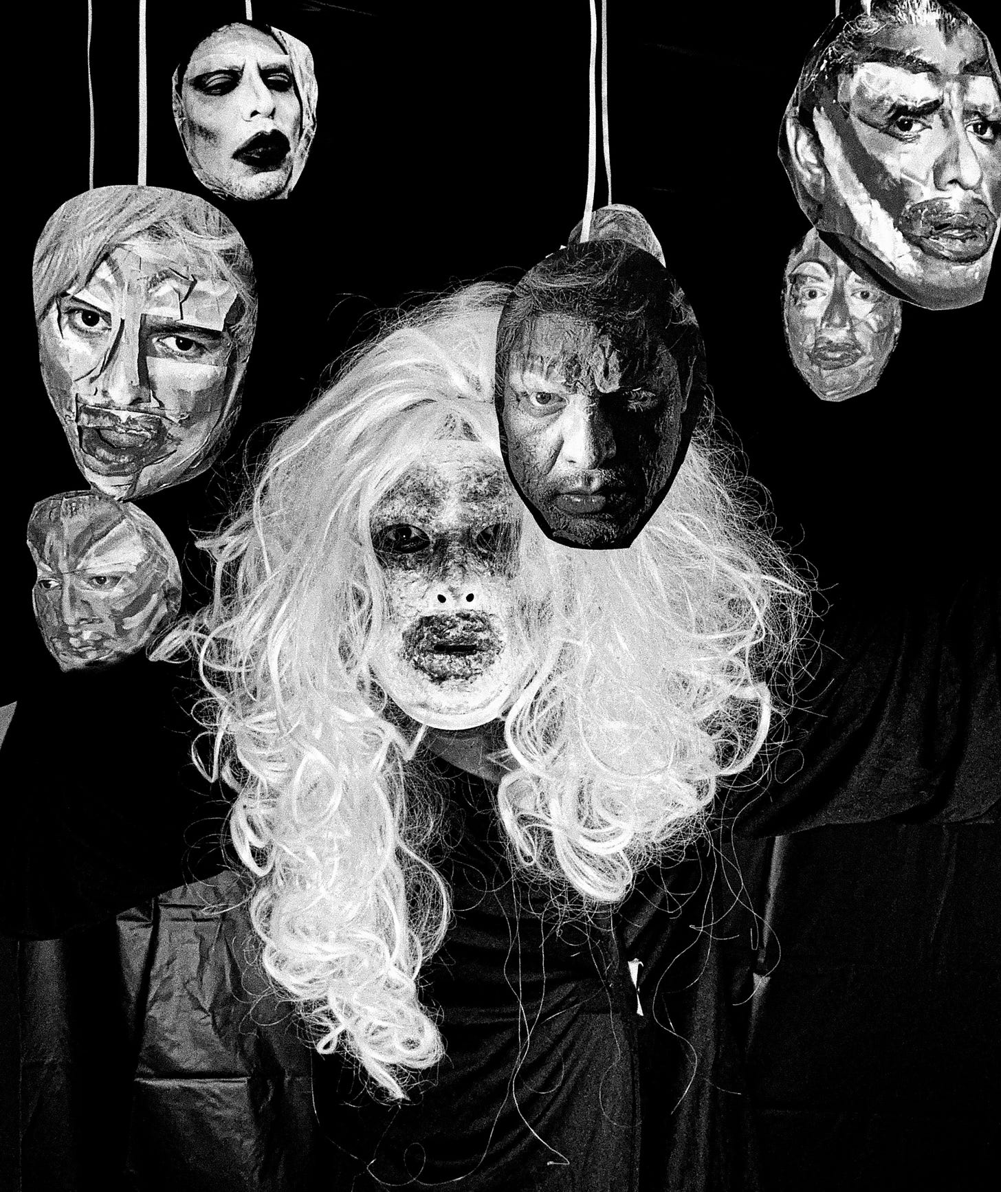 High-contrast, black-and-white photographic portrait of Krystal Lake Carrington. She is face-on, wearing a long silvery curly wig while wearing something black in front of a black background. Her make-up is ghostly white with dark blotchiness around her lips, eyes, and eyebrows. She is staring dead-eyed into the camera amidst six other cut-out Krystal Lake faces that are hanging down around her on white ropes.