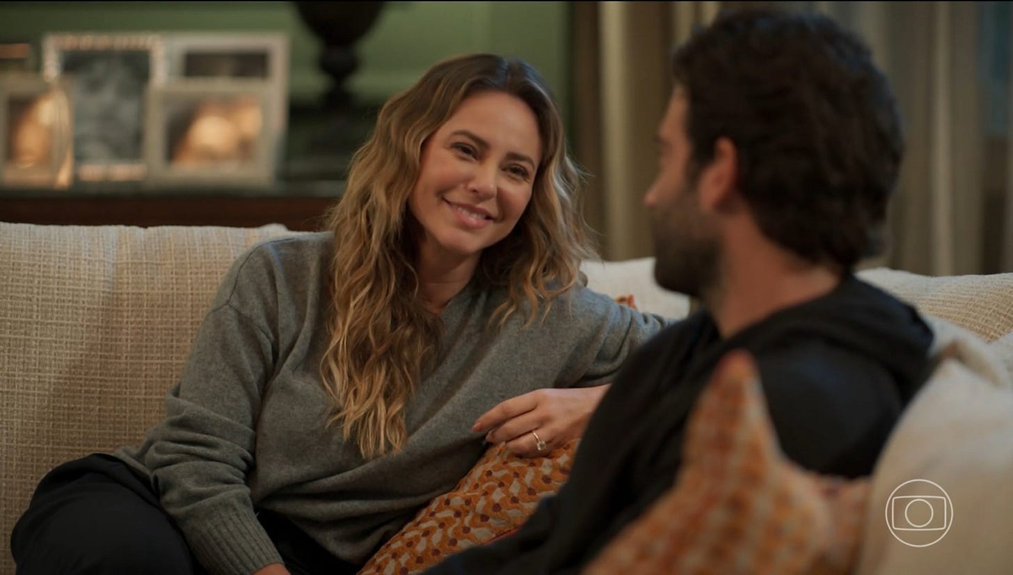 A imagem mostra Heleninha Roitman e Afonso Roitman, personagens da novela Vale Tudo, sentados lado a lado em um sofá, em um momento de intimidade e cumplicidade. Heleninha, interpretada por Paolla Oliveira, está sorrindo carinhosamente para Afonso, vivido por Humberto Carrão, com expressão afetuosa e corpo levemente inclinado em sua direção. Ela veste um suéter cinza e tem cabelos ondulados soltos. Afonso, de costas para a câmera, parece atento e envolvido na conversa. O ambiente é acolhedor, com almofadas e porta-retratos ao fundo, sugerindo uma atmosfera doméstica e familiar.