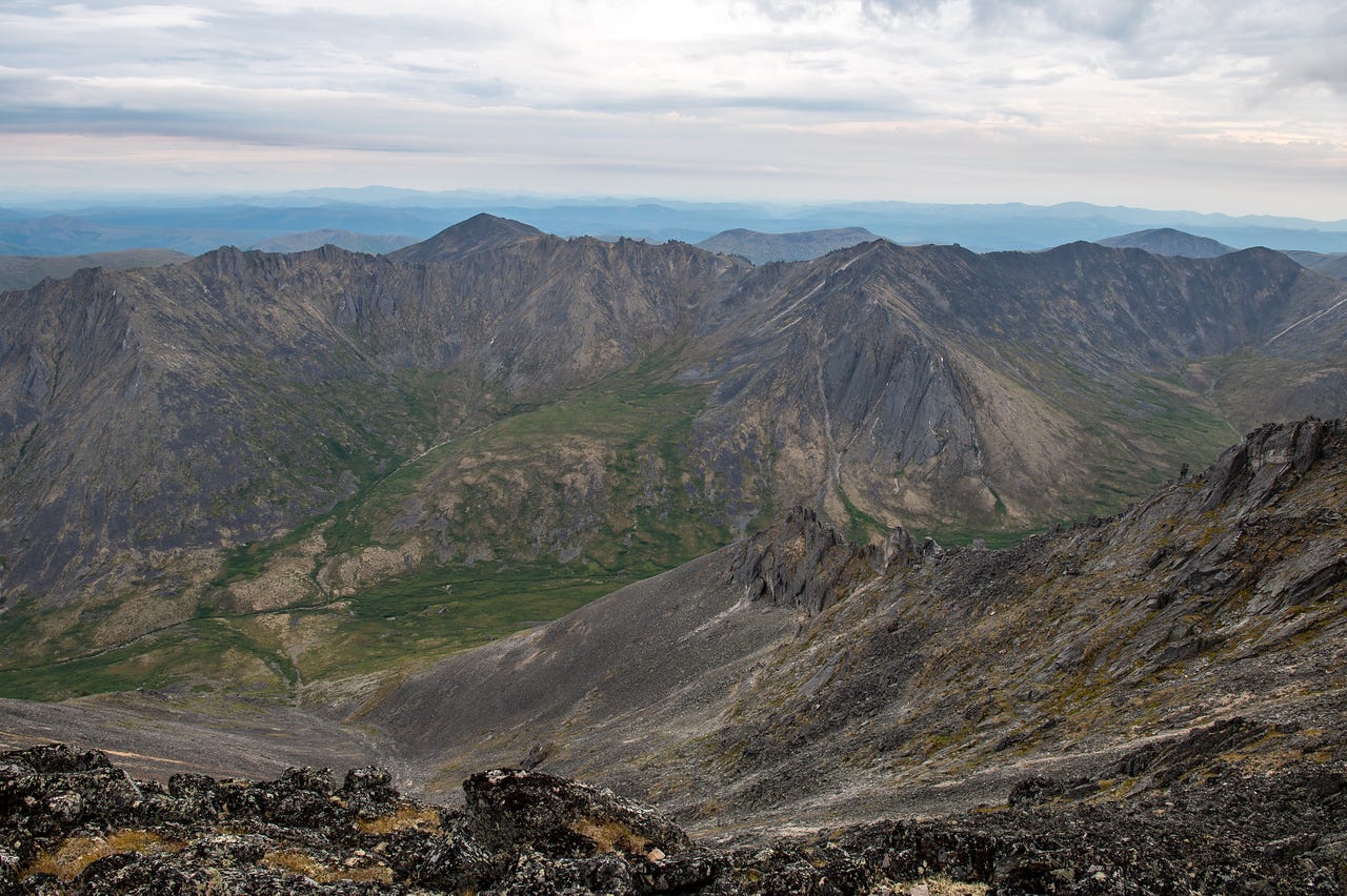A high summit view over row after row of rugged grey-green ridges and deep valleys, a broad tundra basin below and faint blue ranges fading to the horizon under a soft grey sky. Lichen-blotched summit rocks fill the foreground.