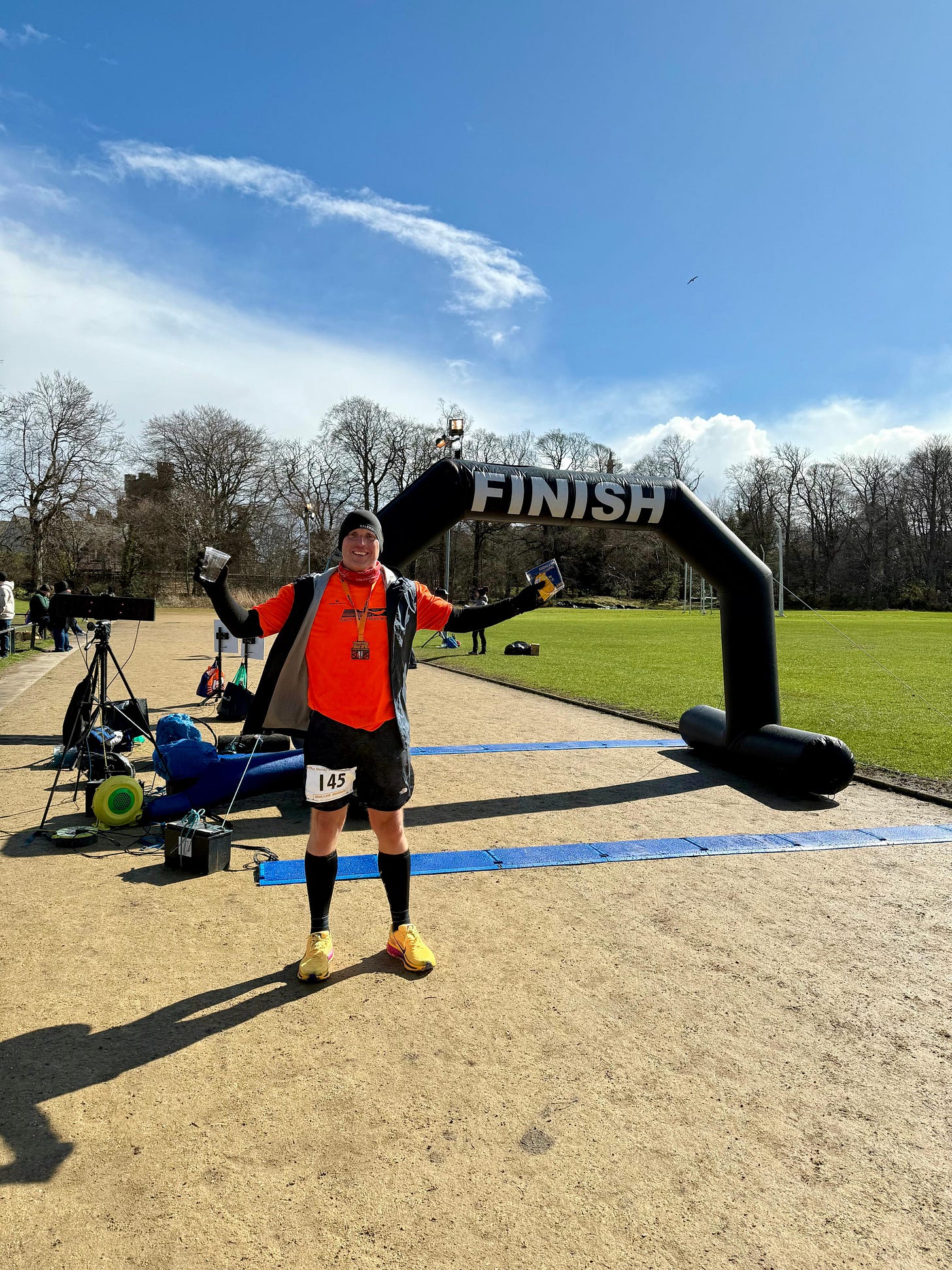 Neil Mapes standing triumphantly under a black inflatable finish line arch at the Moray Marathon. He is smiling broadly with his arms spread wide, wearing a bright orange running shirt, black shorts, a black beanie, and yellow running shoes on a sunny day.