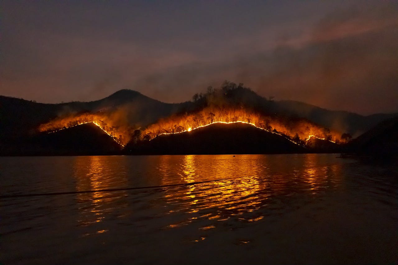 A photo taken at nighttime. There is a lake in the foreground and backdrop featuring hills with a brushfire going across the width of the photo and the light of the fire is reflected on the water. A photo taken at nighttime. There is a lake in the foreground and backdrop featuring hills with a brushfire going across the width of the photo and the light of the fire is reflected on the water.