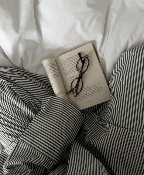 This may contain: an open book laying on top of a bed next to eyeglasses and pillows