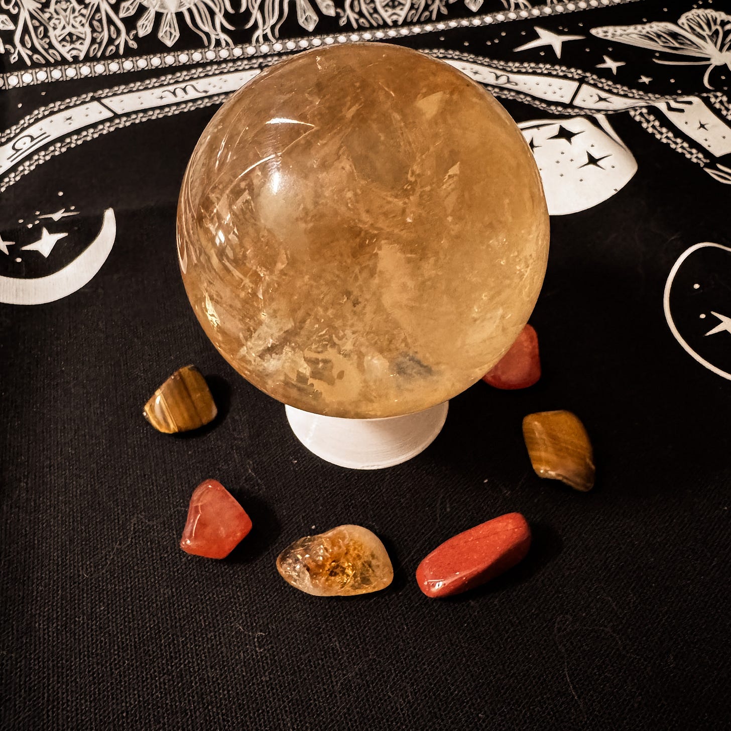 A dark honey-coloured crystal sphere sits on a white pedestal, atop a black altar cloth decorated with white lunar symbols. Around the pedestal is a circle of red- and orange-coloured tumble stones. The crystals featured are carnelian, citrine, red jasper, tiger’s eye, and honey calcite.