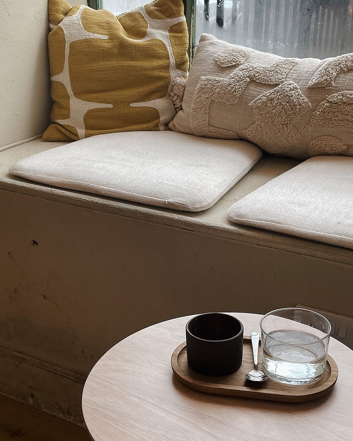 Image 1: Cosy window seating with cushions, a small wooden table holding a cup of coffee and a glass of water. / Image 2: Scandi-inspired seating with high stools, wooden furniture, and a vase of dried flowers in soft beige and yellow tones.