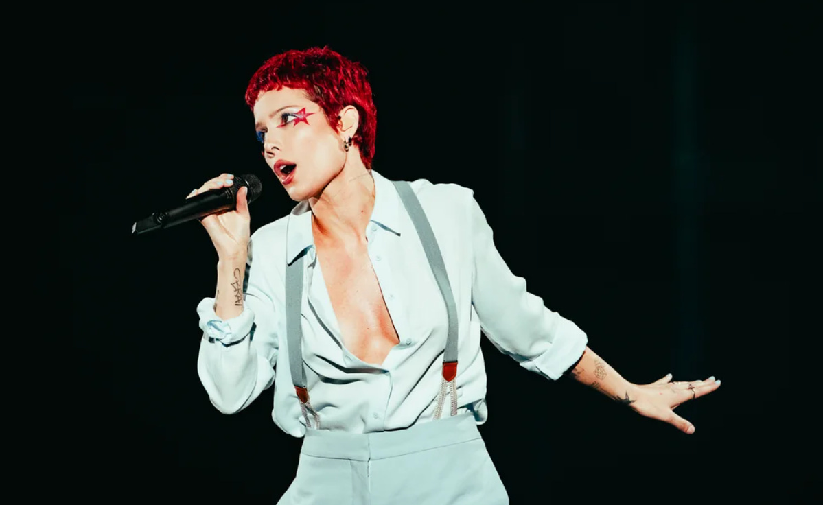 Halsey, with red short hair and a blue two piece outfit with suspenders, holding a black microphone.