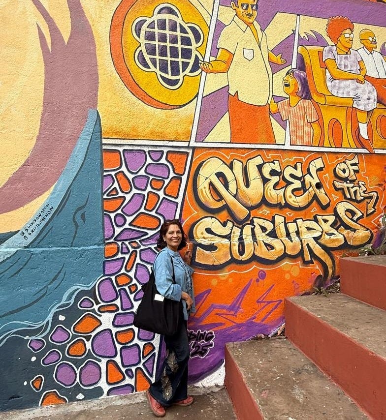 Anu Arora standing in front of a bright, colorful mural that reads Queen of the Suburbs