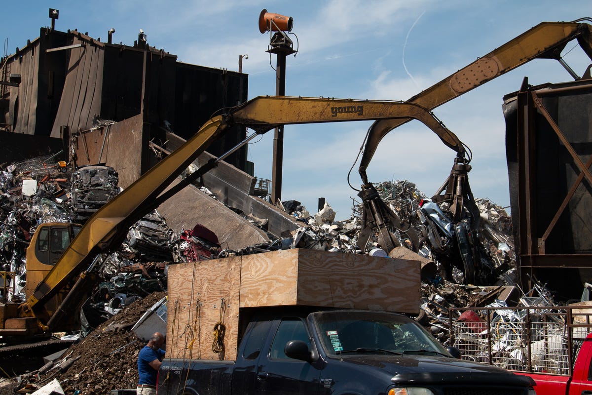 Excavator claws hovering above a pile of scrap metal, with a truck and person in blue shirt in foreground. Excavator claws hovering above a pile of scrap metal, with a truck and person in blue shirt in foreground.