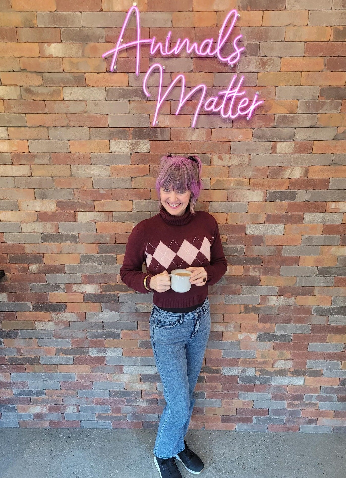 Sadie Freely standing against a brick wall under a pink neon sign reading “Animals Matter,” wearing a purple turtleneck and holding a coffee cup.