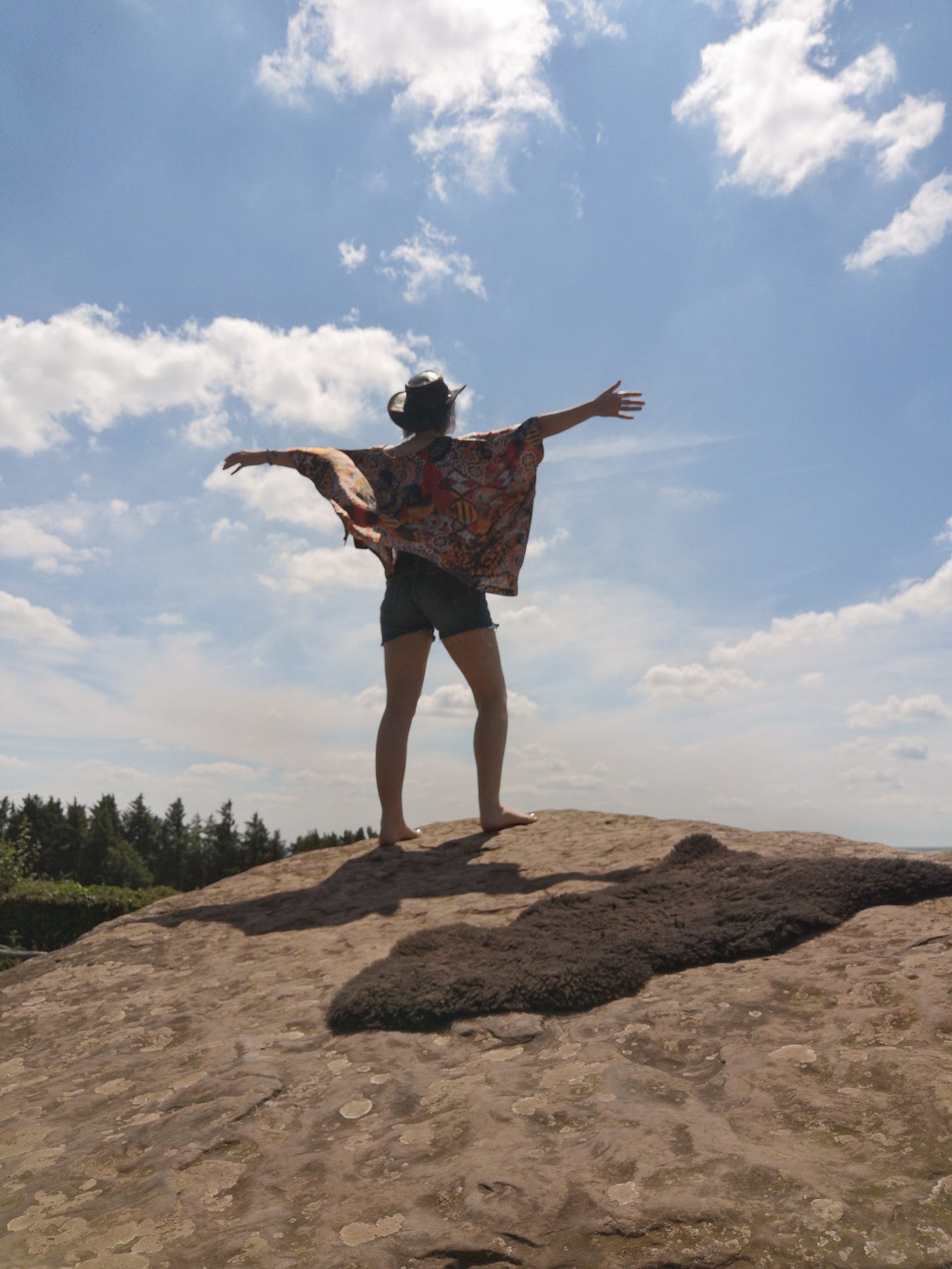 Layne faces away from the camera, standing on a rock with arms stretched. The sky is blue with light clouds
