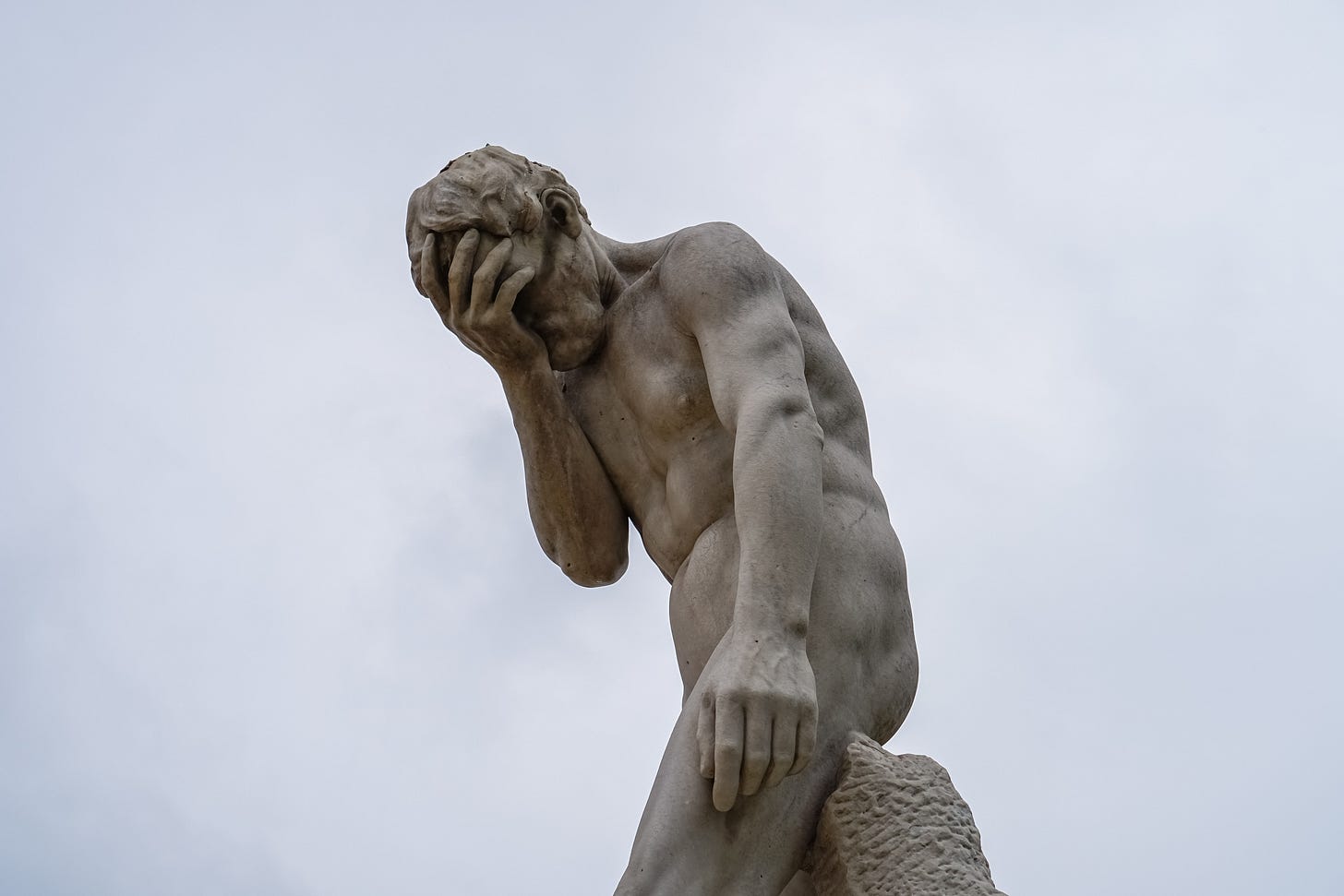 Von leicht schräg unten fotografierte, graue Steinstatue eines nackten Mannes, der sich mit dem Po an eine Art Mauer anlehnt. Die rechte Hand ist vors Gesicht gehalten (Facepalm), der Kopf ist gebeugt, der linke Arm hängt seitlich nah am Körper herunter. Im Hintergrund grauer Himmel. Von leicht schräg unten fotografierte, graue Steinstatue eines nackten Mannes, der sich mit dem Po an eine Art Mauer anlehnt. Die rechte Hand ist vors Gesicht gehalten (Facepalm), der Kopf ist gebeugt, der linke Arm hängt seitlich nah am Körper herunter. Im Hintergrund grauer Himmel.